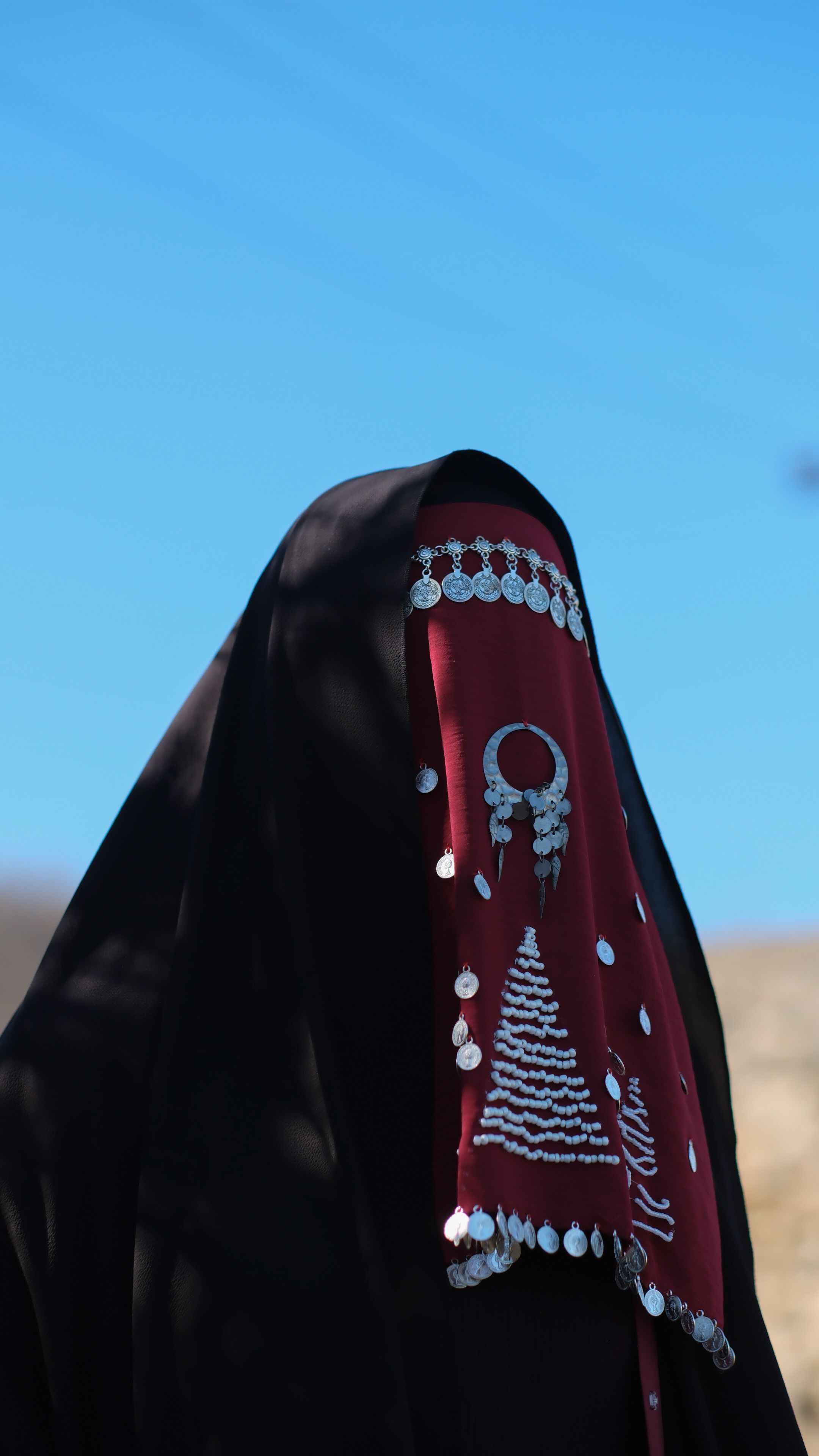 A mysterious figure in traditional clothing with a red and black veil adorned with jewelry against a clear blue sky.