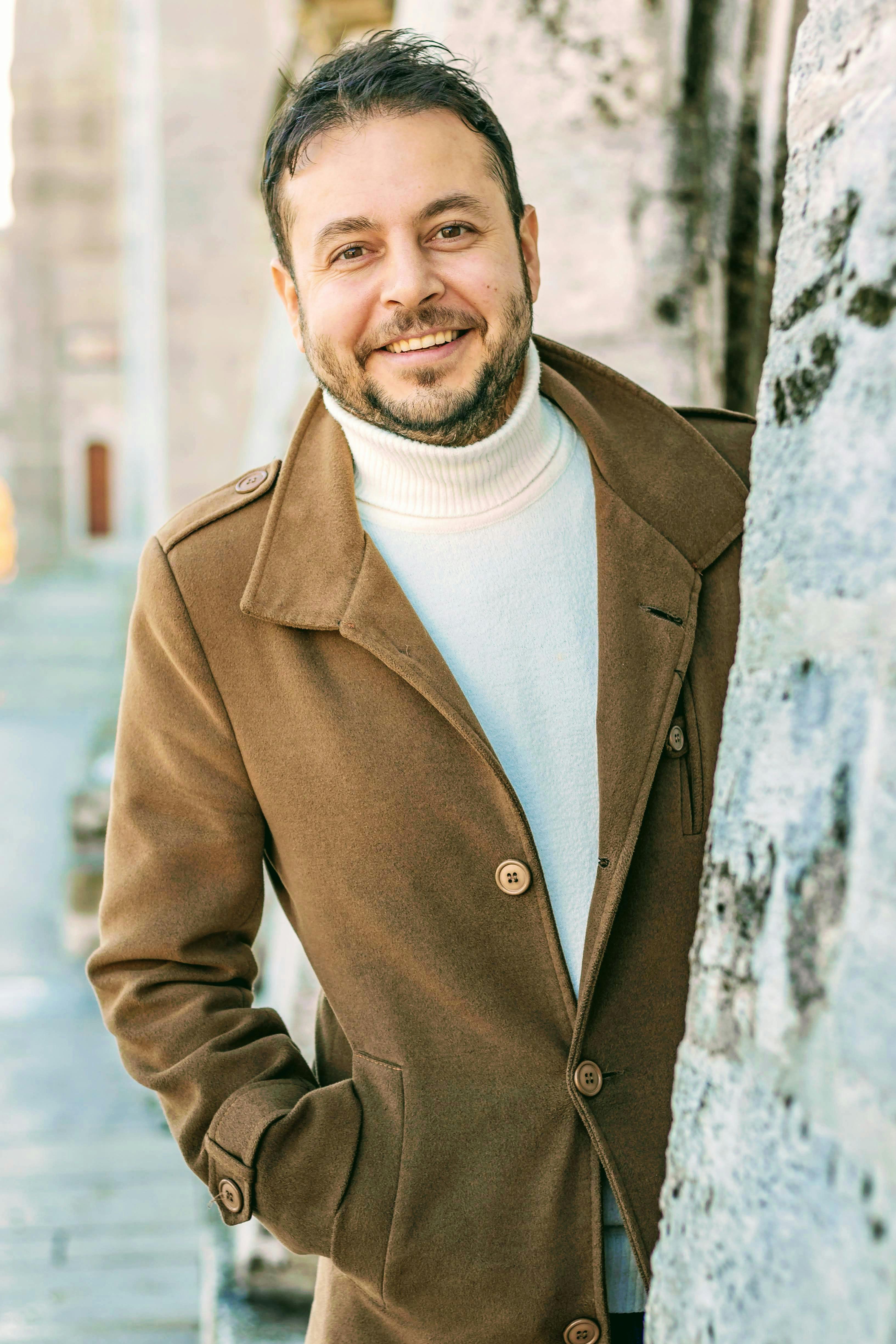 Portrait of a smiling man in a brown coat leaning against a stone wall during sunset.