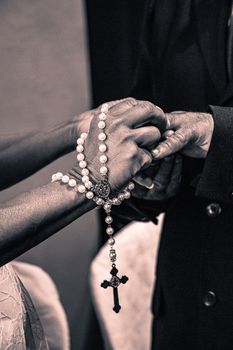 Close-up of bride and groom holding hands with rosary beads during wedding ceremony.