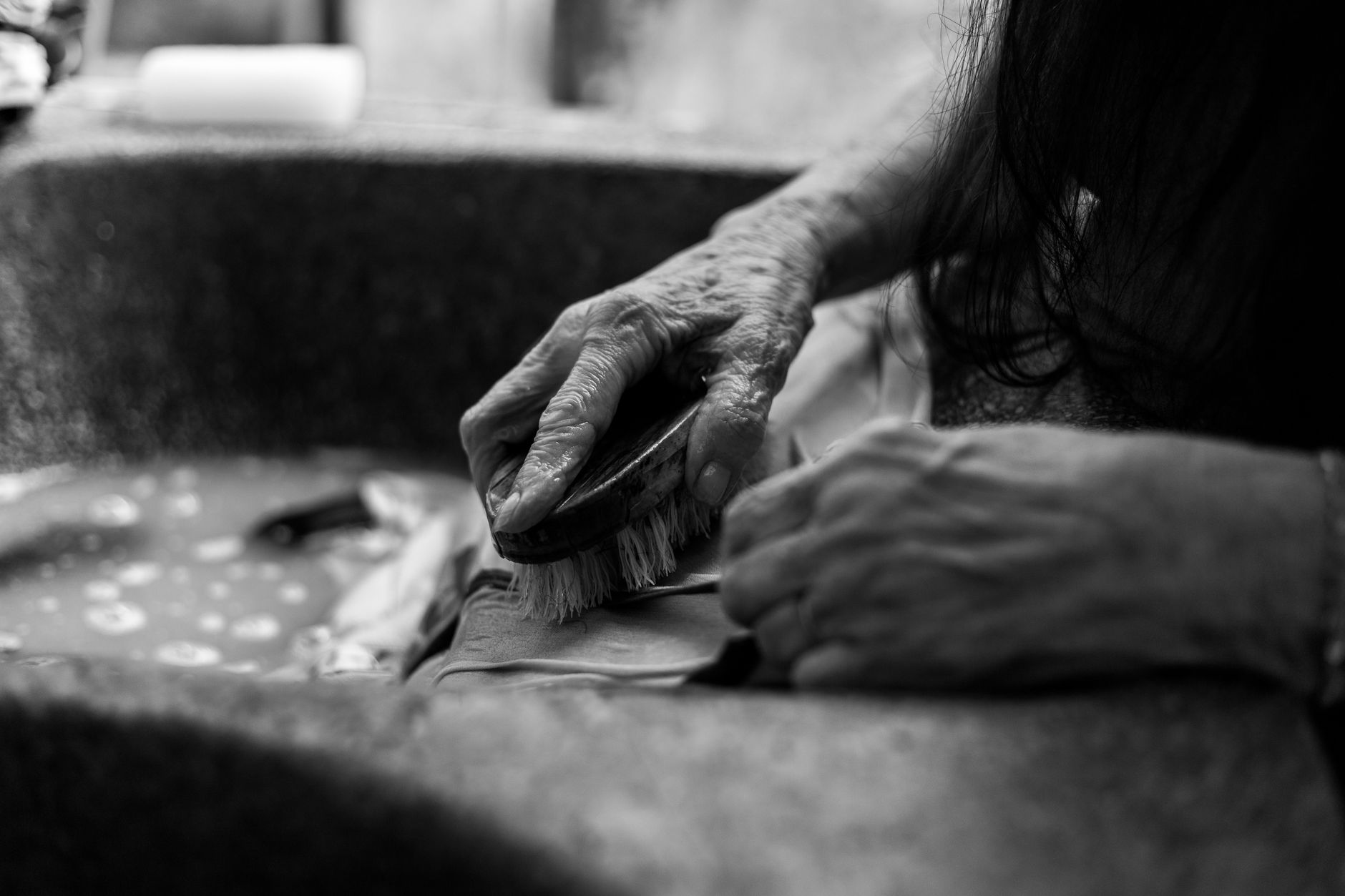 An elderly woman handwashing clothes using a brush in a rustic rural setting, captured in black and white.