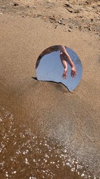 Artistic shot of hands reflecting on a mirror placed on a sandy beach with gentle waves.
