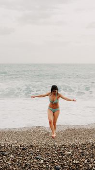 A woman in a bikini enjoys a beach day by the ocean, standing on the pebbled shore.