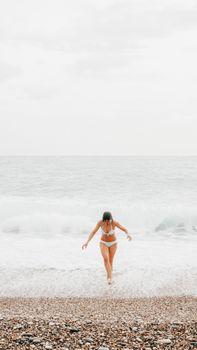 A woman in a bikini walks along a rocky beach with waves crashing around her.