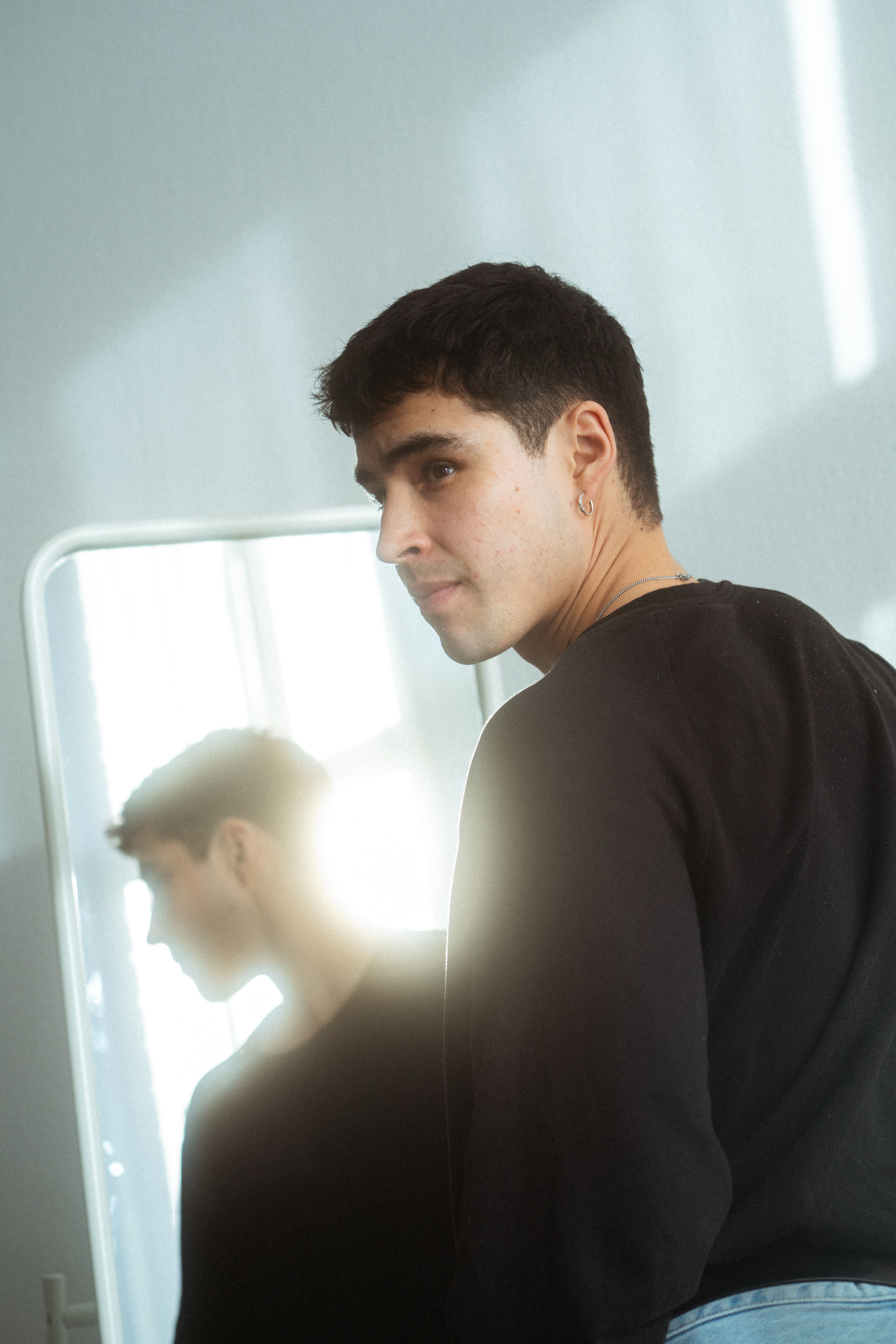 A man in a black sweater looks at his reflection in a mirror, capturing natural light.