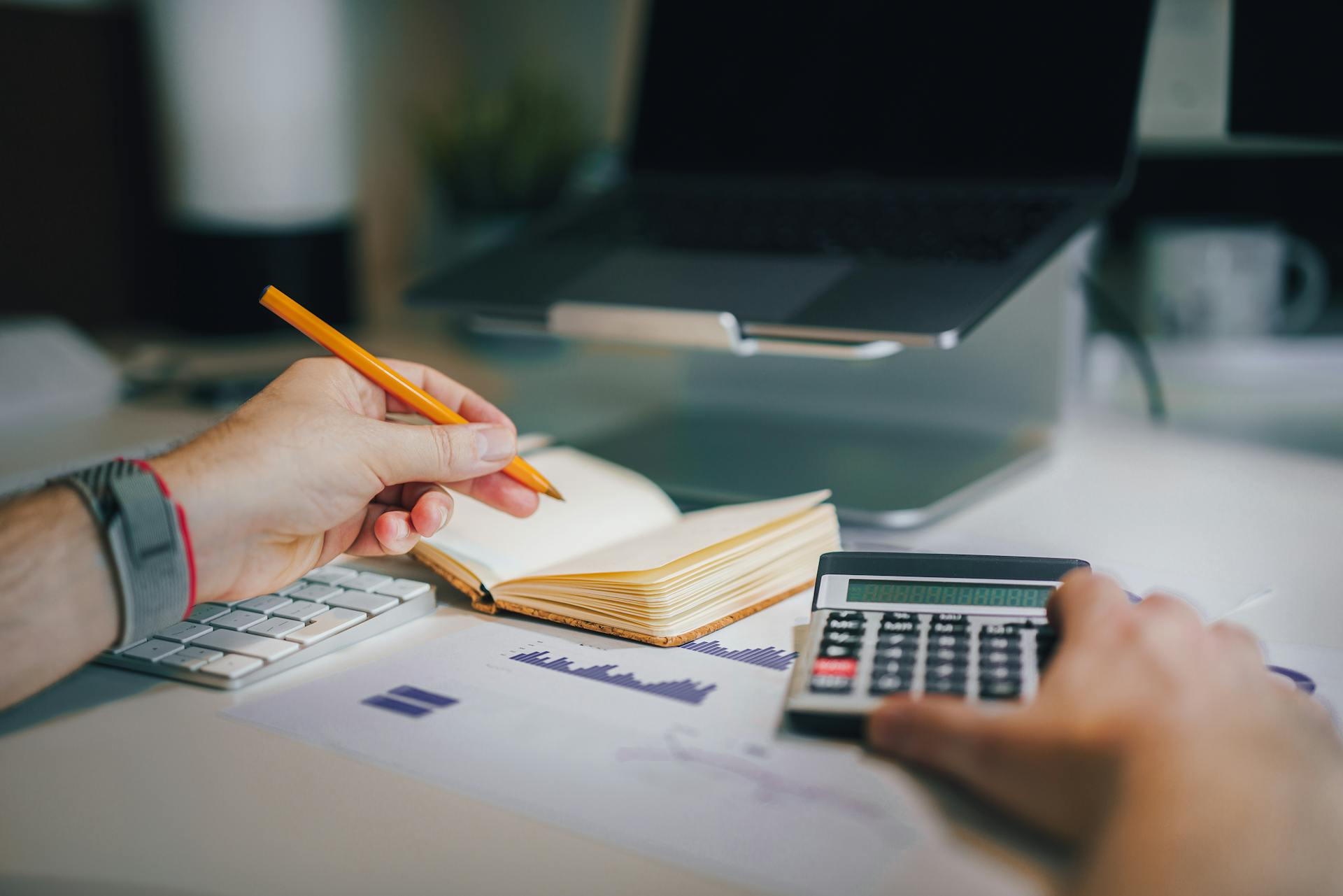 Hands using calculator and taking notes on desk with laptop, showcasing business analytics.