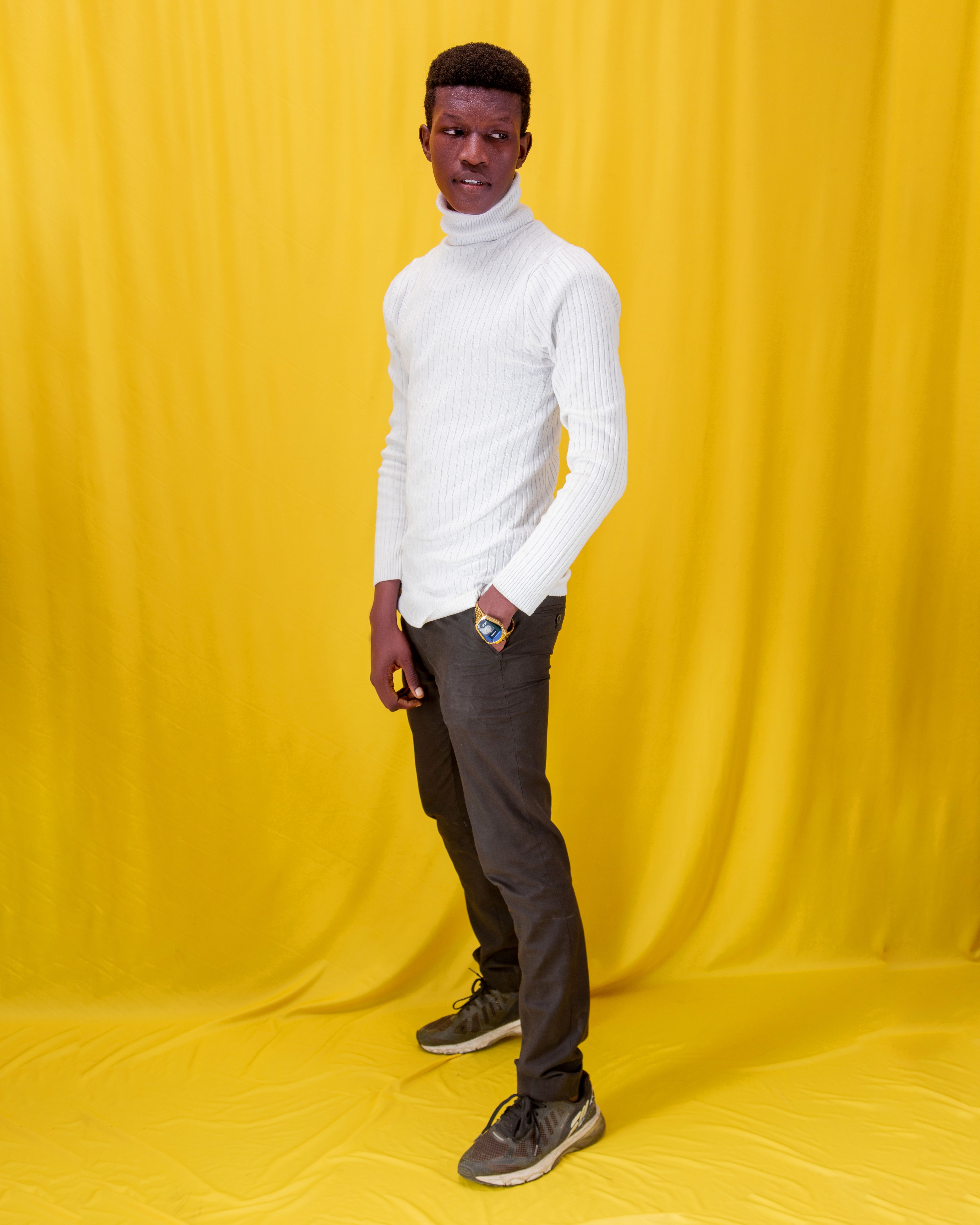 Fashion portrait of a stylish man posing in a white turtleneck and black pants against a yellow background.