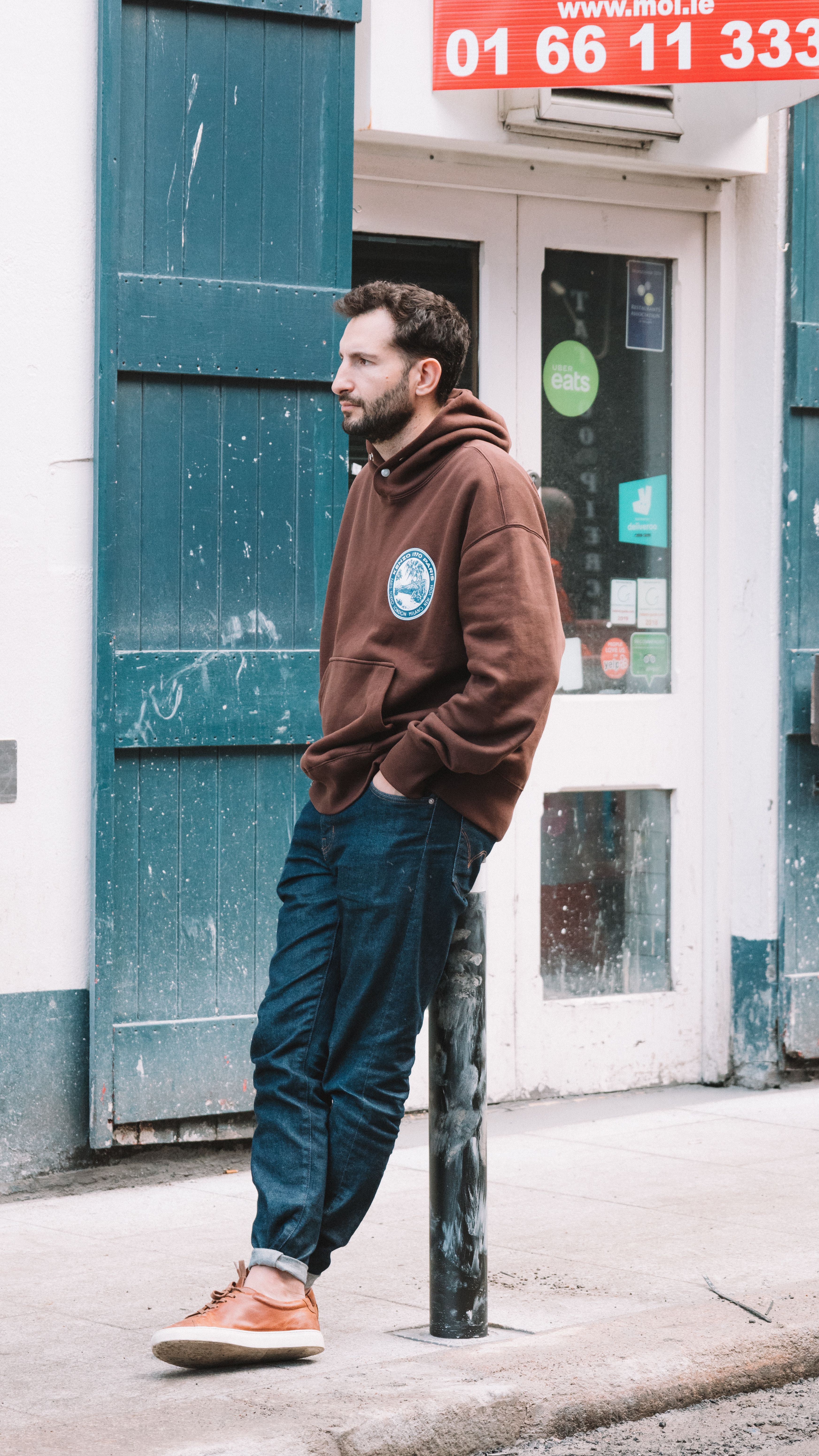 Man in brown hoodie leaning against post on urban sidewalk.