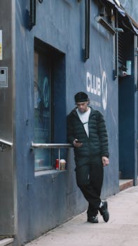 Man in black jacket leaning on a blue wall with signage, checking phone, urban setting.