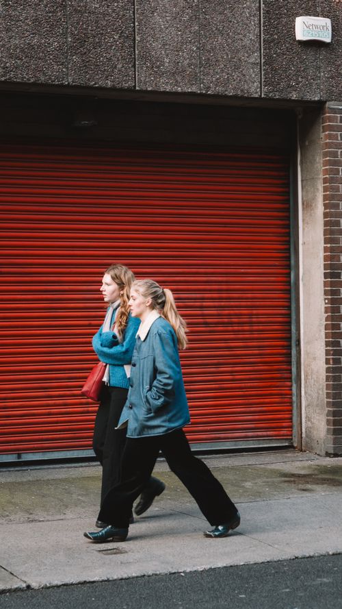 Free Two women walking in front of a red shutter in an urban setting, showcasing fall fashion. Stock Photo