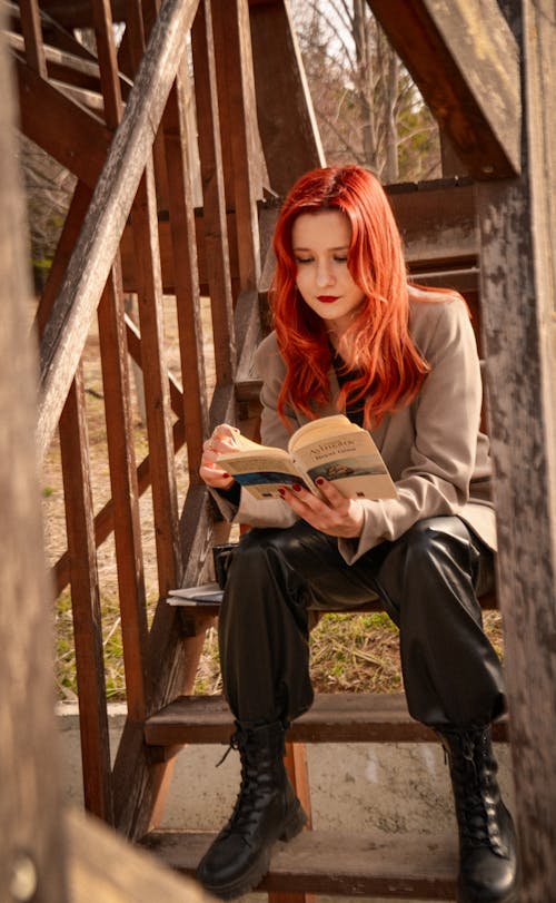 A woman with red hair is sitting on a wooden staircase reading a book