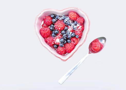 Heart-shaped bowl filled with raspberries and blueberries, accompanied by a spoon on a white background.