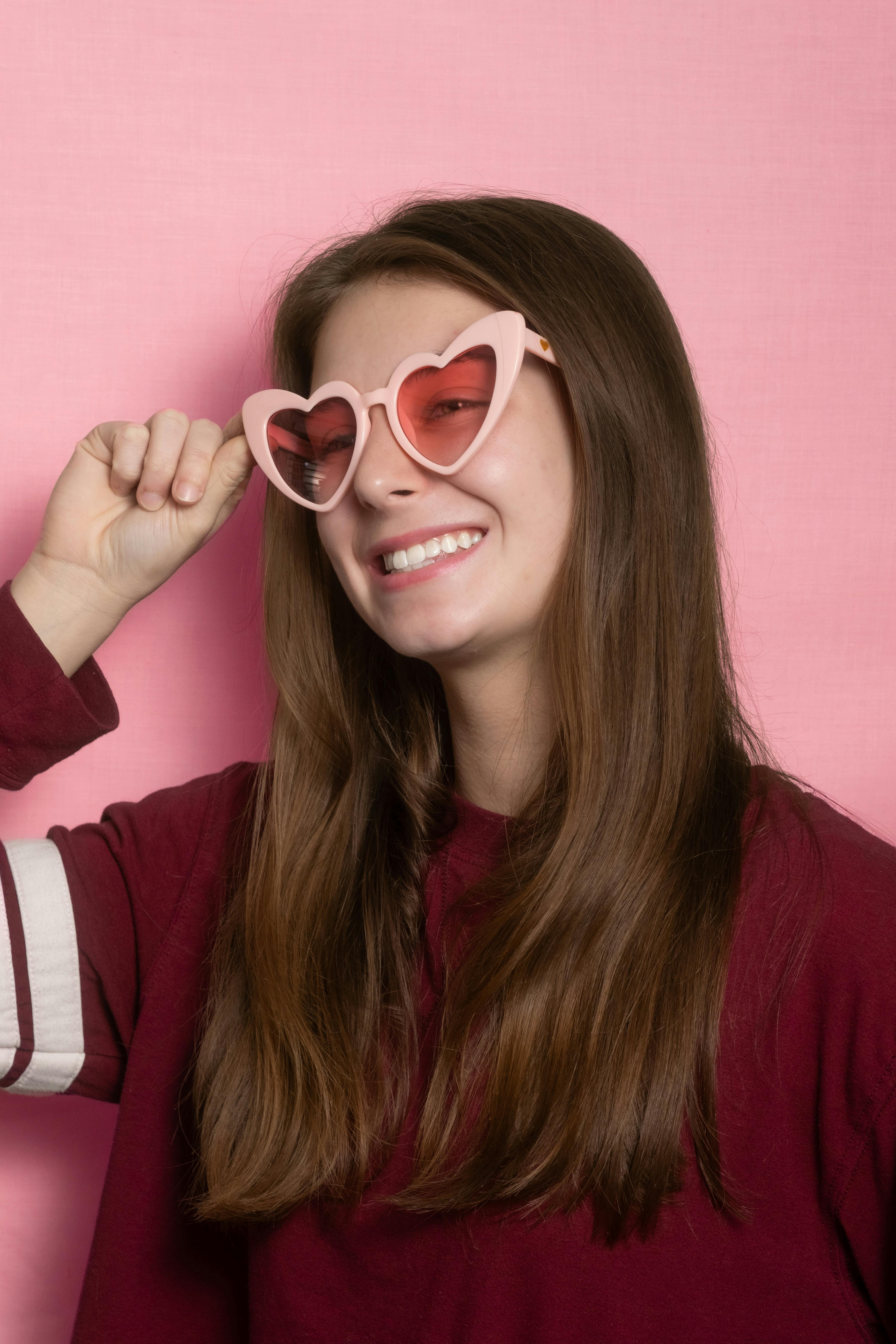 Cheerful young woman in heart-shaped sunglasses posing against a pink background.