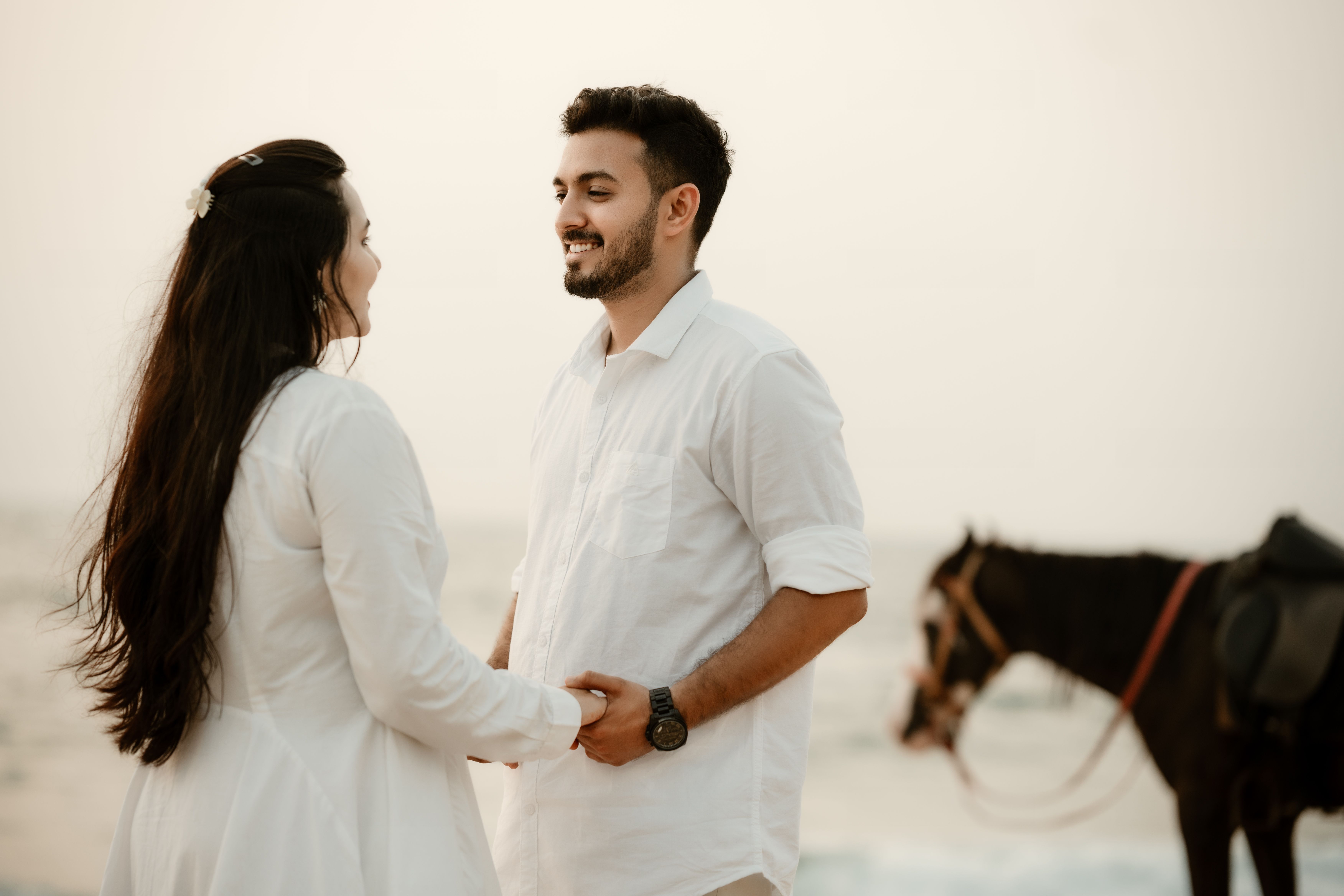 Couple dressed in white enjoying a romantic moment on a beach with a horse in the background at sunset.