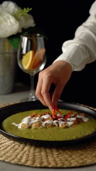 Hand garnishing broccoli soup with red peppers, creamy topping, elegant plating.