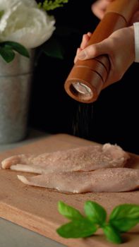 Close-up of seasoning raw chicken fillets with a pepper grinder on a wooden board.