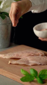 Hand seasoning raw chicken fillets on wooden cutting board with fresh basil. Culinary preparation image.