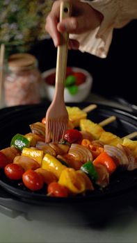 Hand holding brush applying sauce on colorful vegetable skewers on a grill.