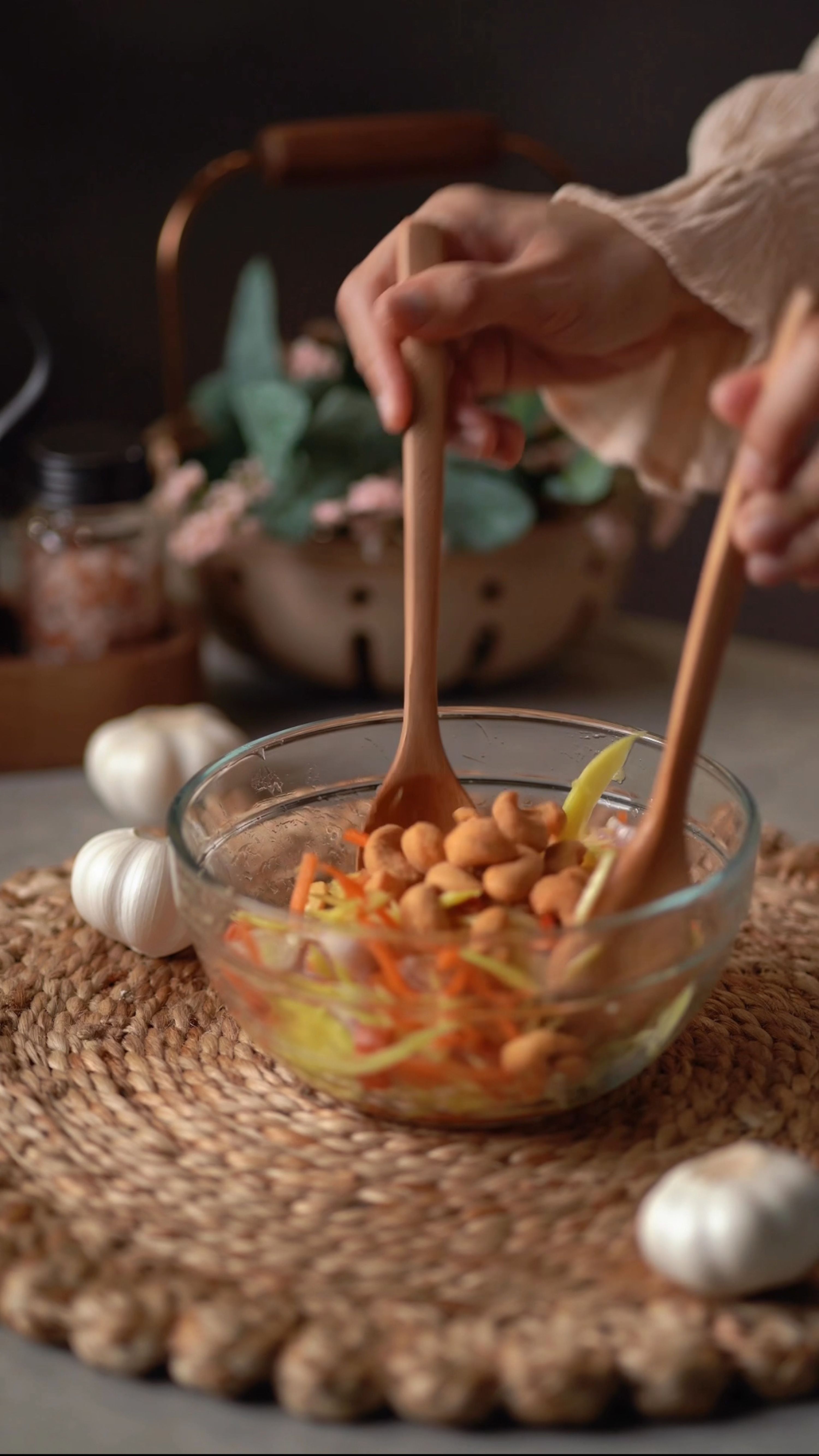 Close-up of hands using wooden spoons to mix a fresh vegetable salad in a glass bowl.
