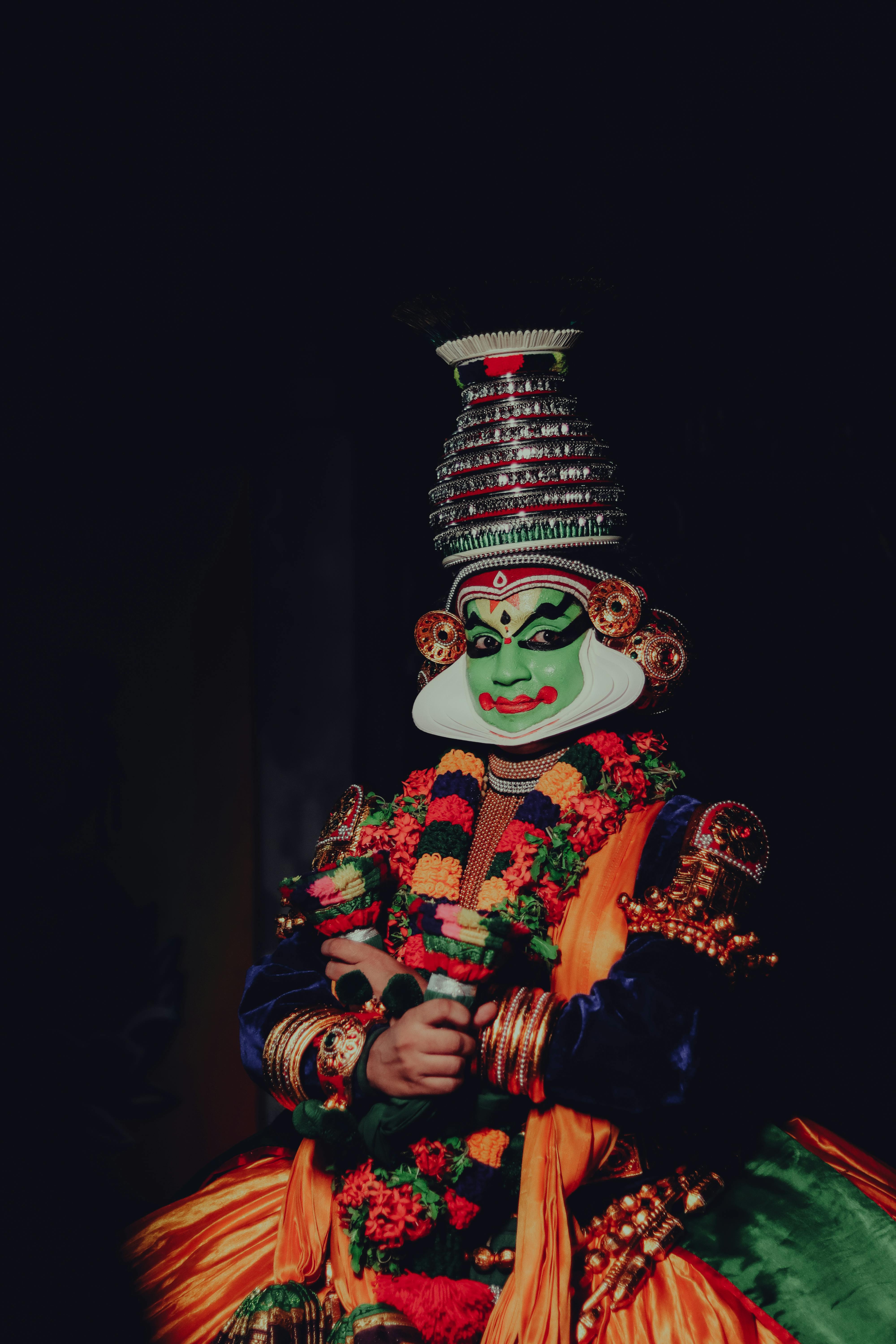 Free Vibrant portrait of a Kathakali performer in traditional attire showcasing intricate makeup and costume against a dark background. Stock Photo