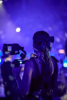 Back view of a female videographer filming an event with vibrant lighting in a nightclub setting.