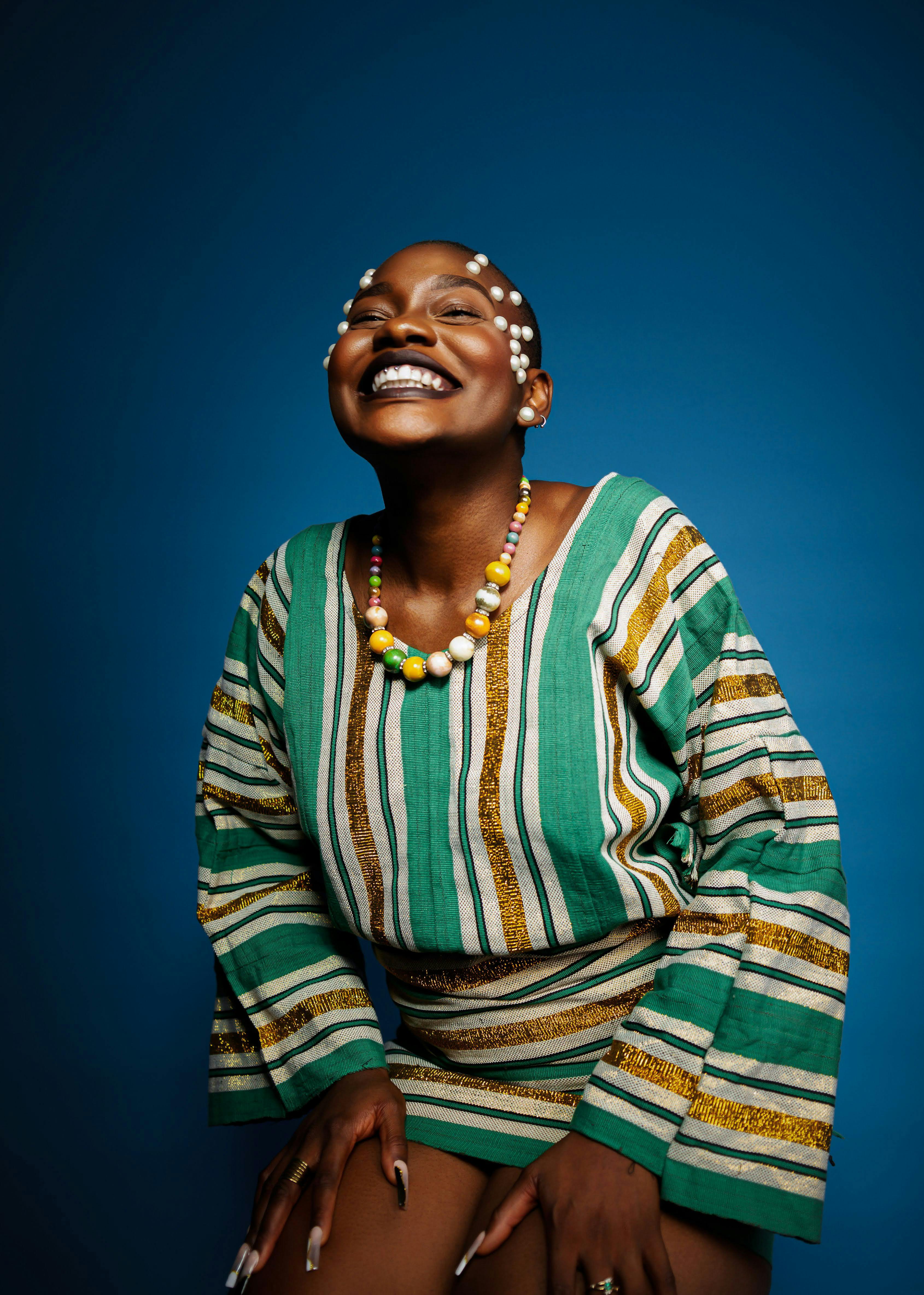 A smiling African woman poses in traditional striped clothing and beaded necklace on a blue background.