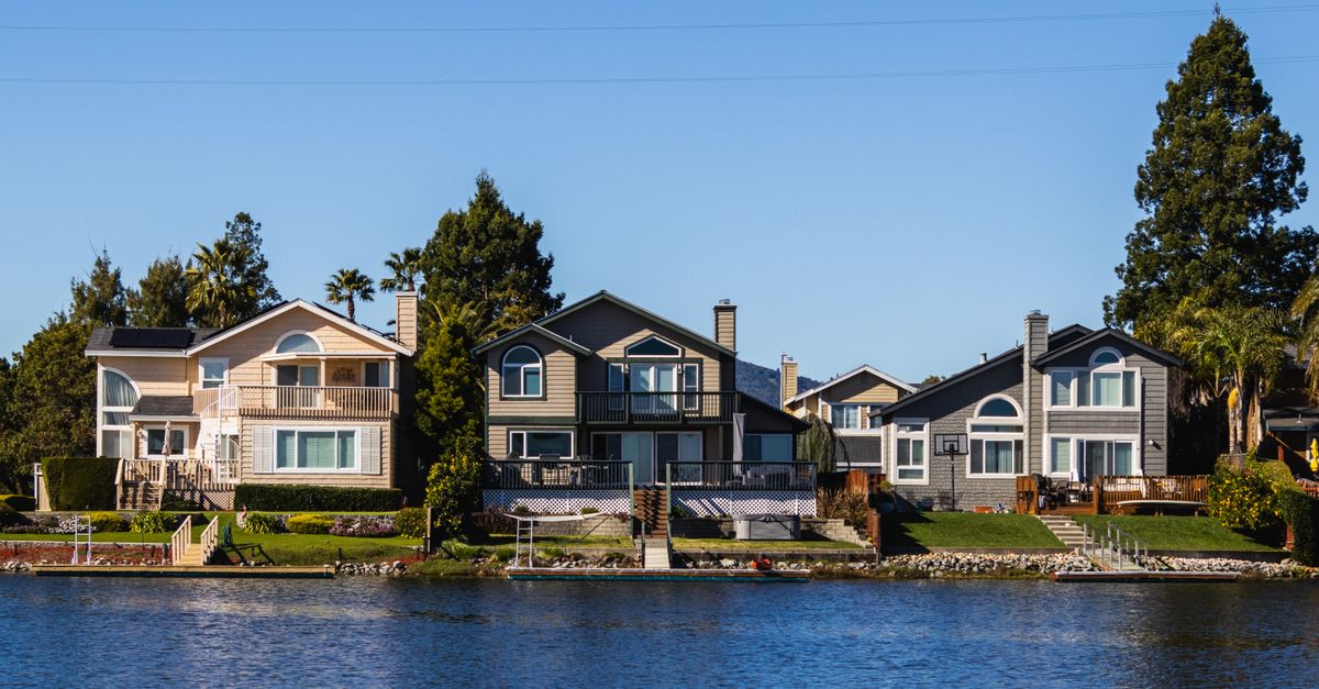 Charming lakeside houses reflect on calm waters under a clear blue sky.