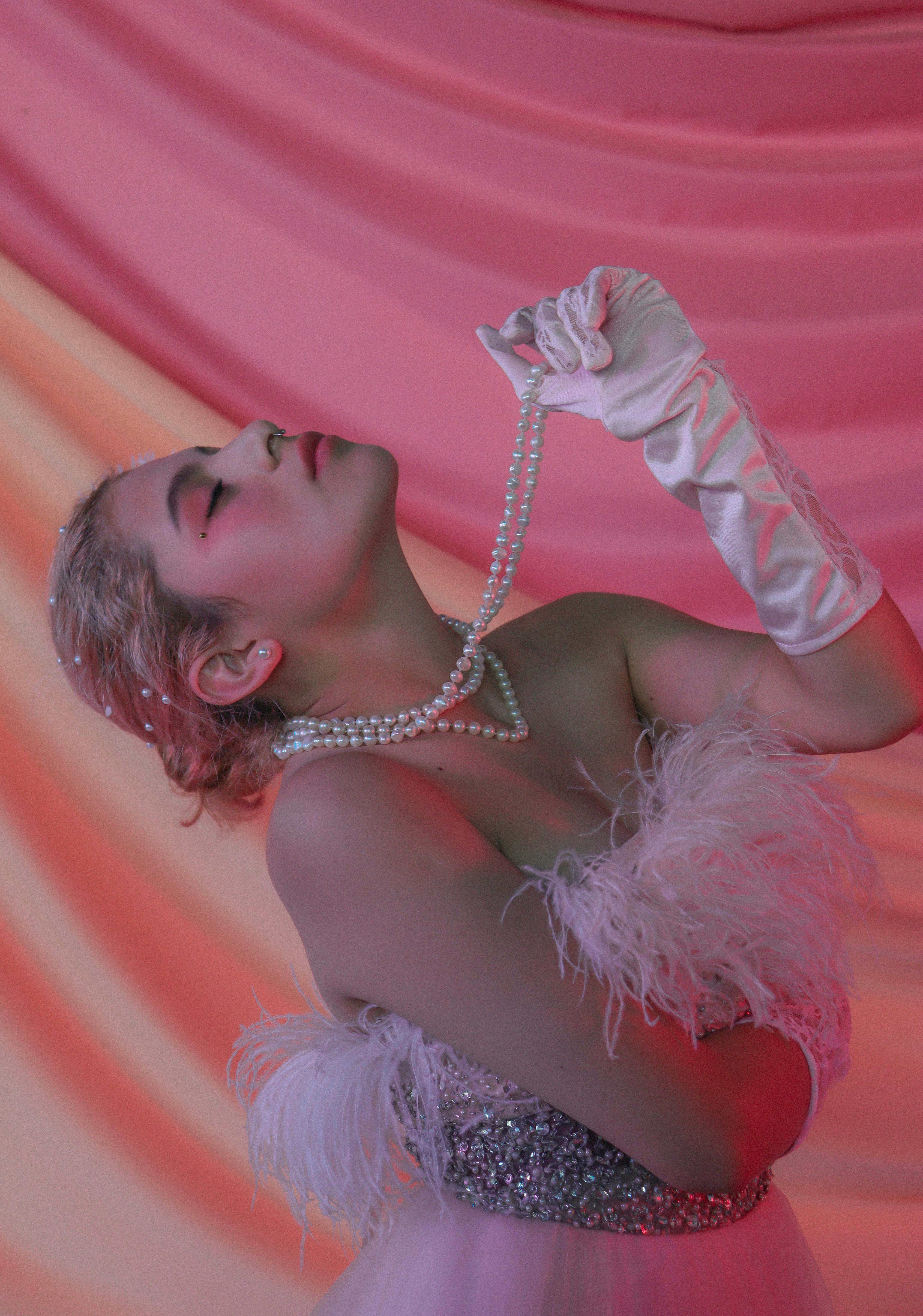 A glamorous studio portrait featuring a woman with pearls and silk gloves against a pink draped background.