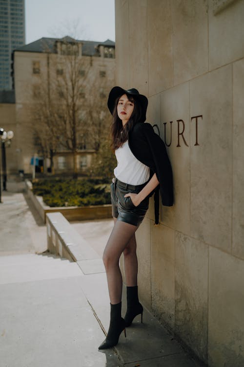A woman in black hat and leather shorts leaning against a wall