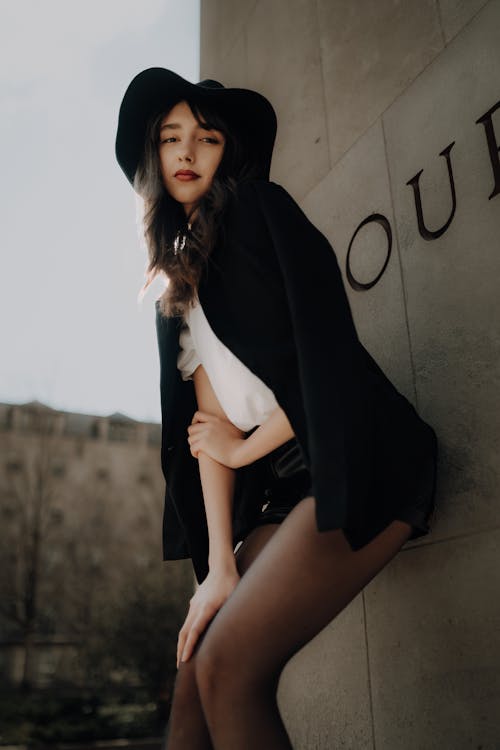 A woman in black hat and stockings posing near a wall