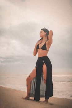 A woman in a black bikini poses gracefully on a beach under a cloudy sky.