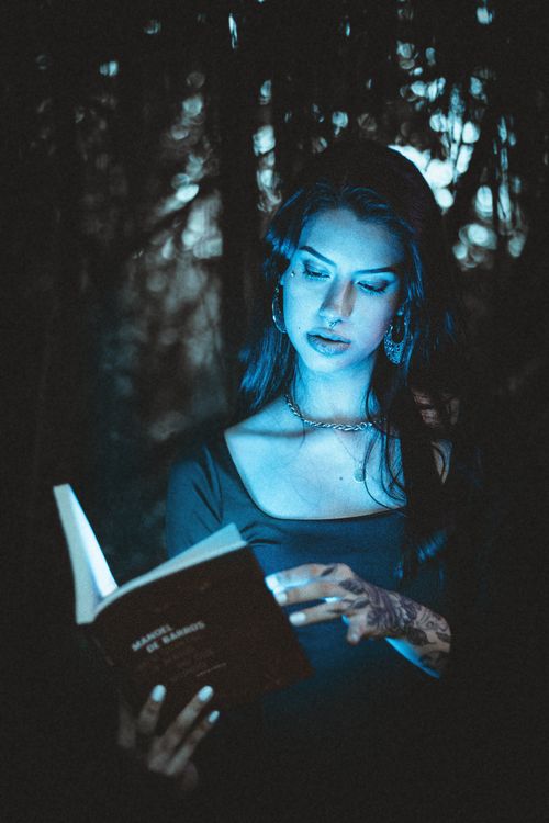 Free A woman with tattoos reads a book in a dark forest, illuminated by blue light. Stock Photo
