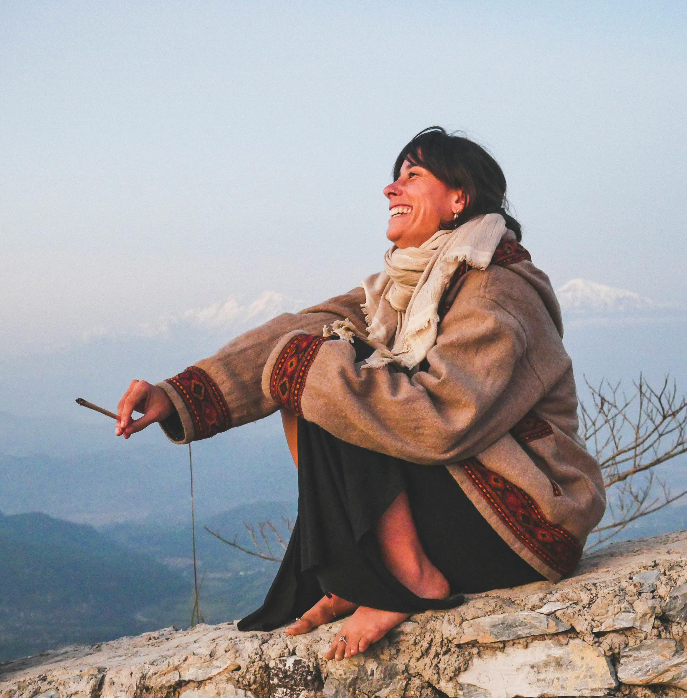A woman sits barefoot on a rock, smiling and enjoying the mountain view at sunset.