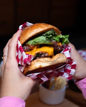 Delicious double cheeseburger with lettuce and meat, wrapped in checkered paper, held by hands.