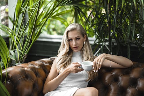 Young Woman in a White Dress Sitting on a Brown Leather Couch with a Cup of Coffee
