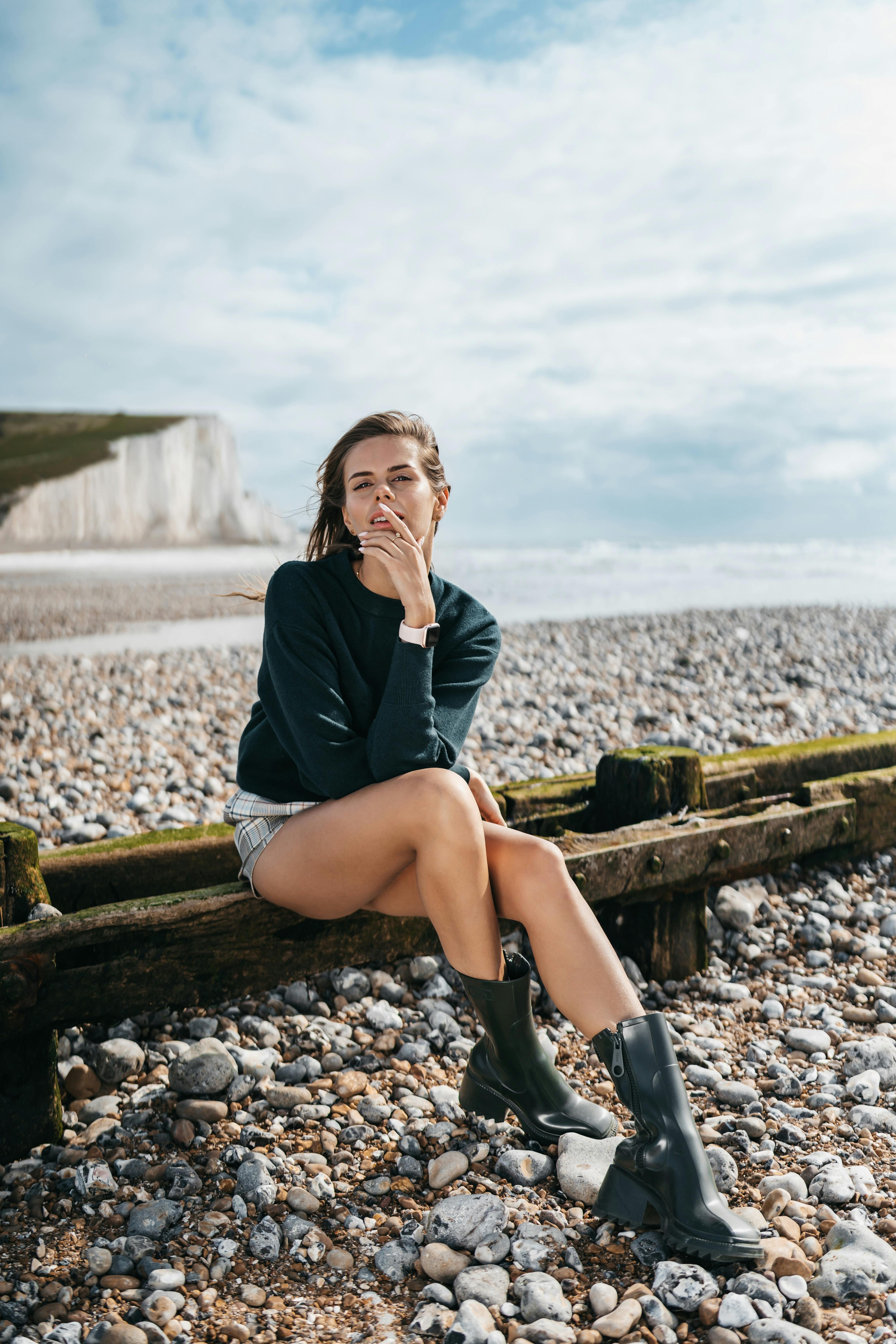 Fashionable woman posing on a pebble beach in stylish boots, with a dramatic coastal backdrop.