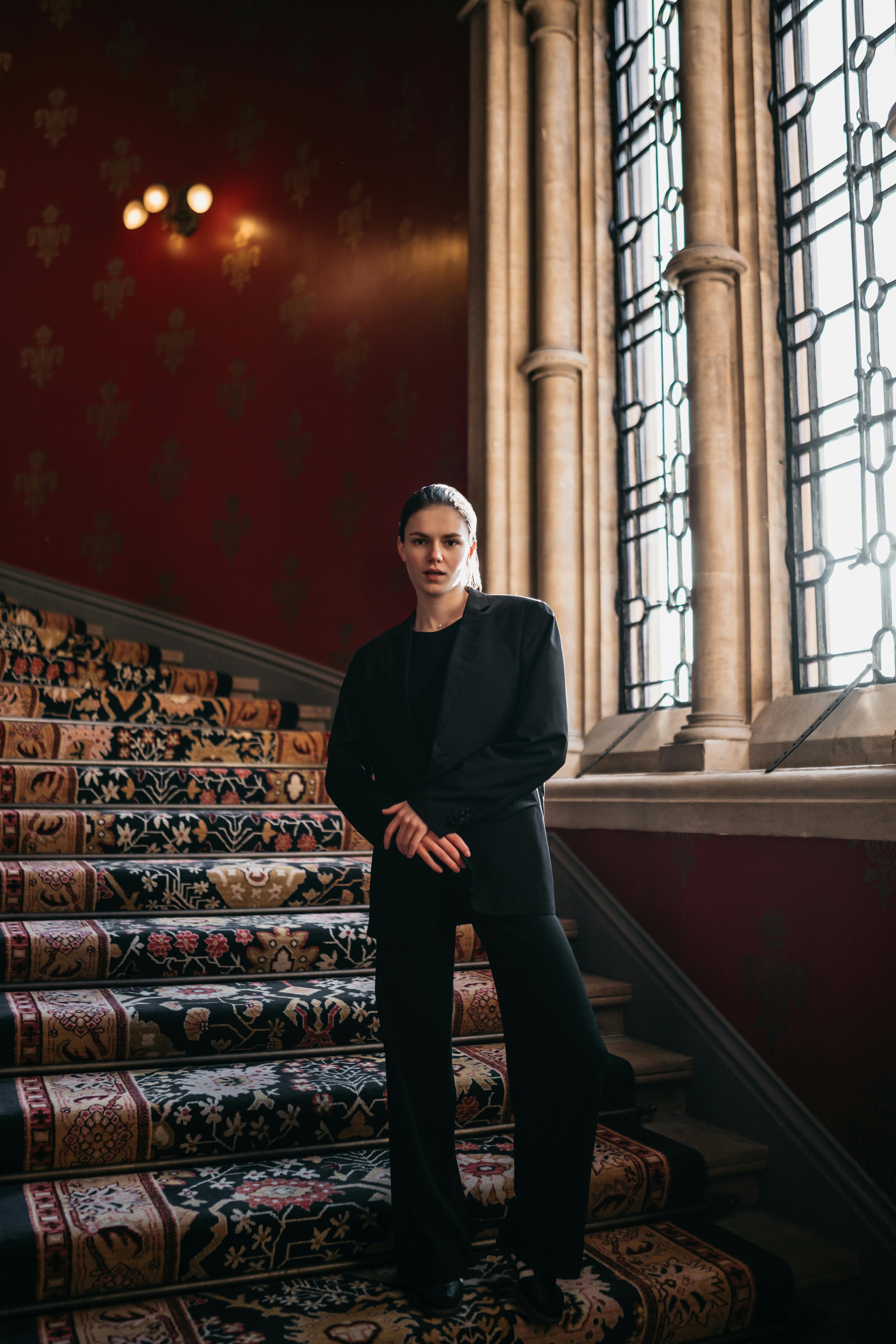 Stylish woman in a suit poses on a classic, ornate staircase with natural light streaming through a large window.
