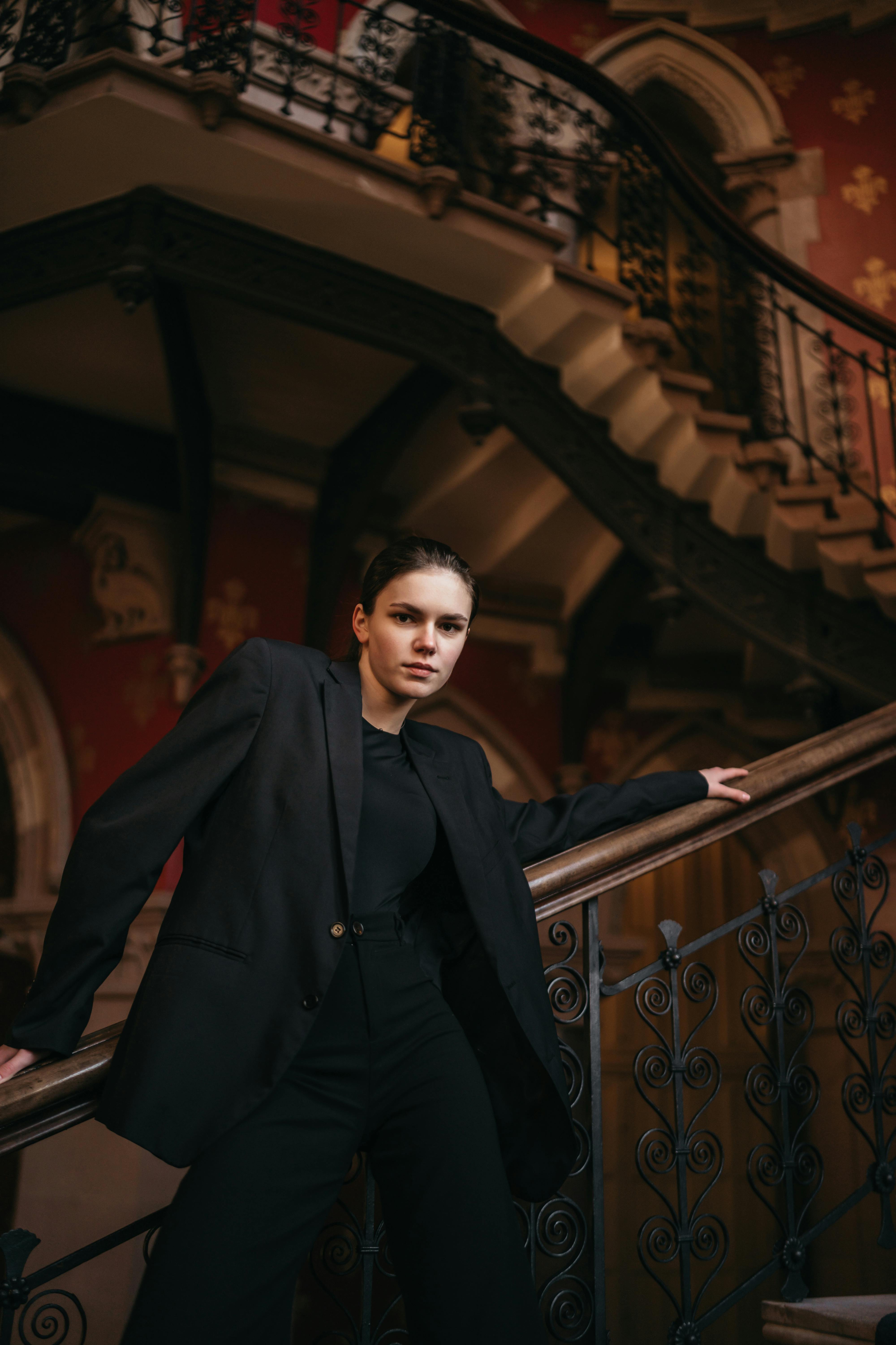 Sophisticated woman in a black suit posing on a grand staircase in an ornate indoor setting.