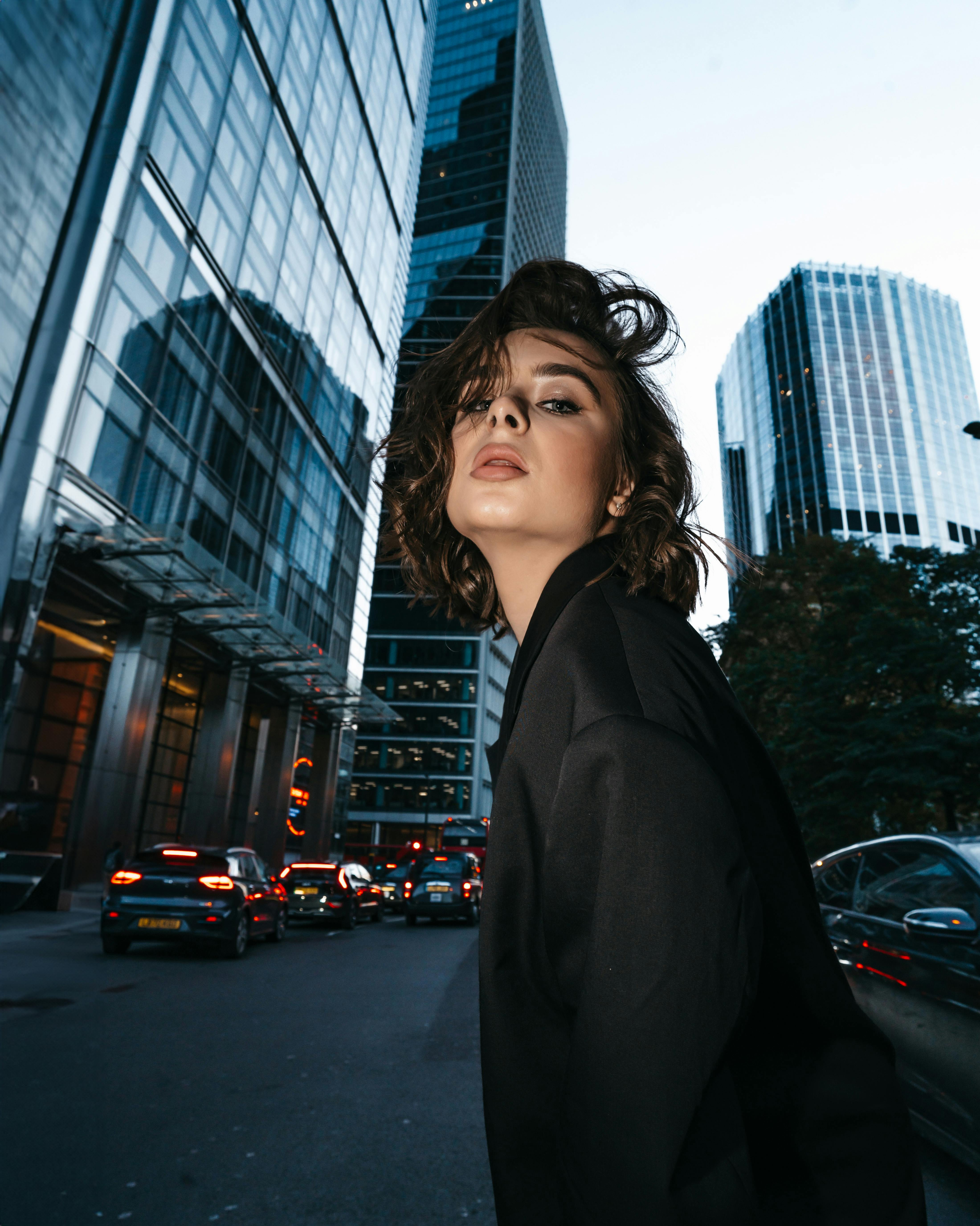 Fashionable woman in black jacket posing against urban skyscrapers at dusk, showcasing modern city life.