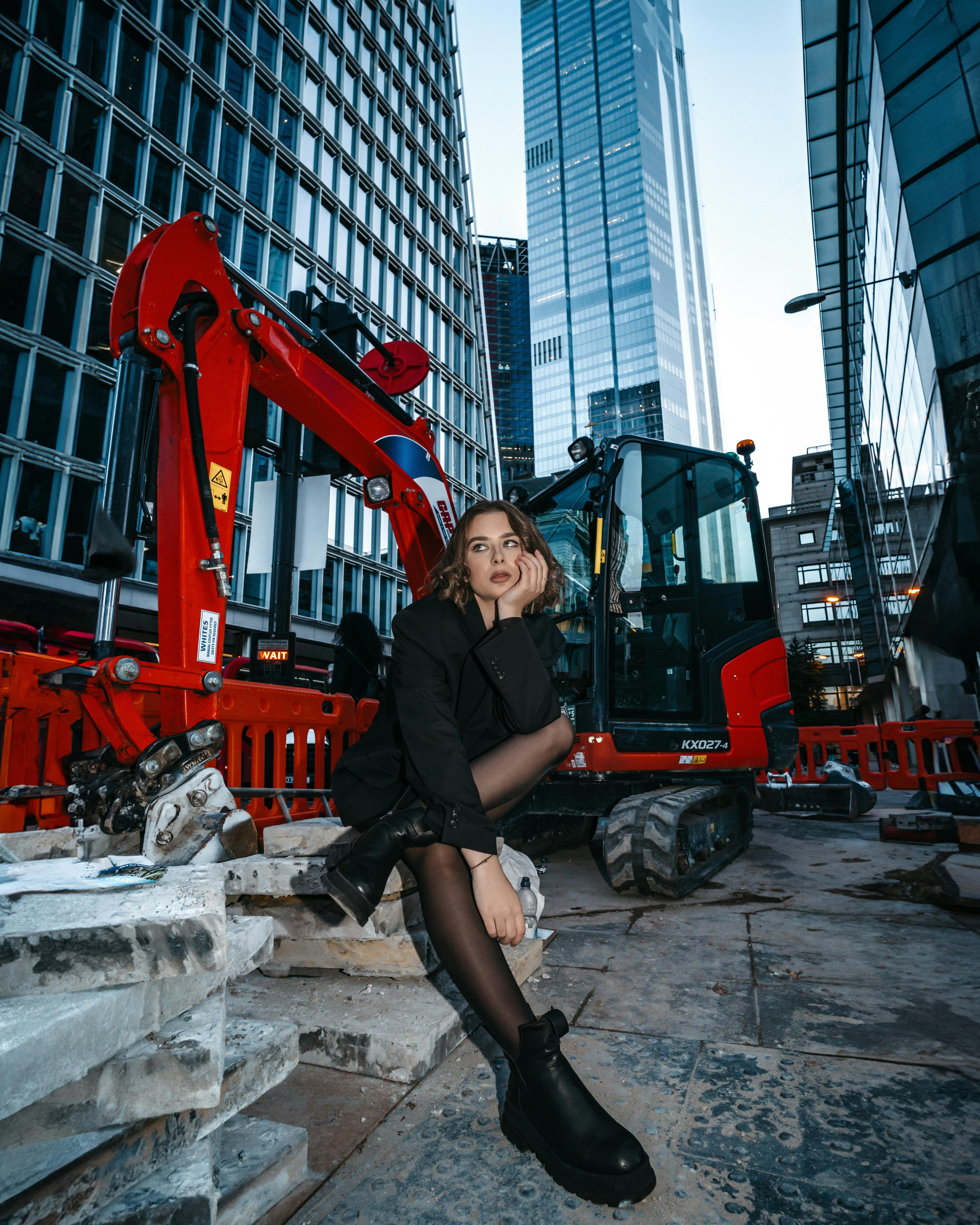 City fashion photo featuring a woman in black outfit sitting by a modern construction site.