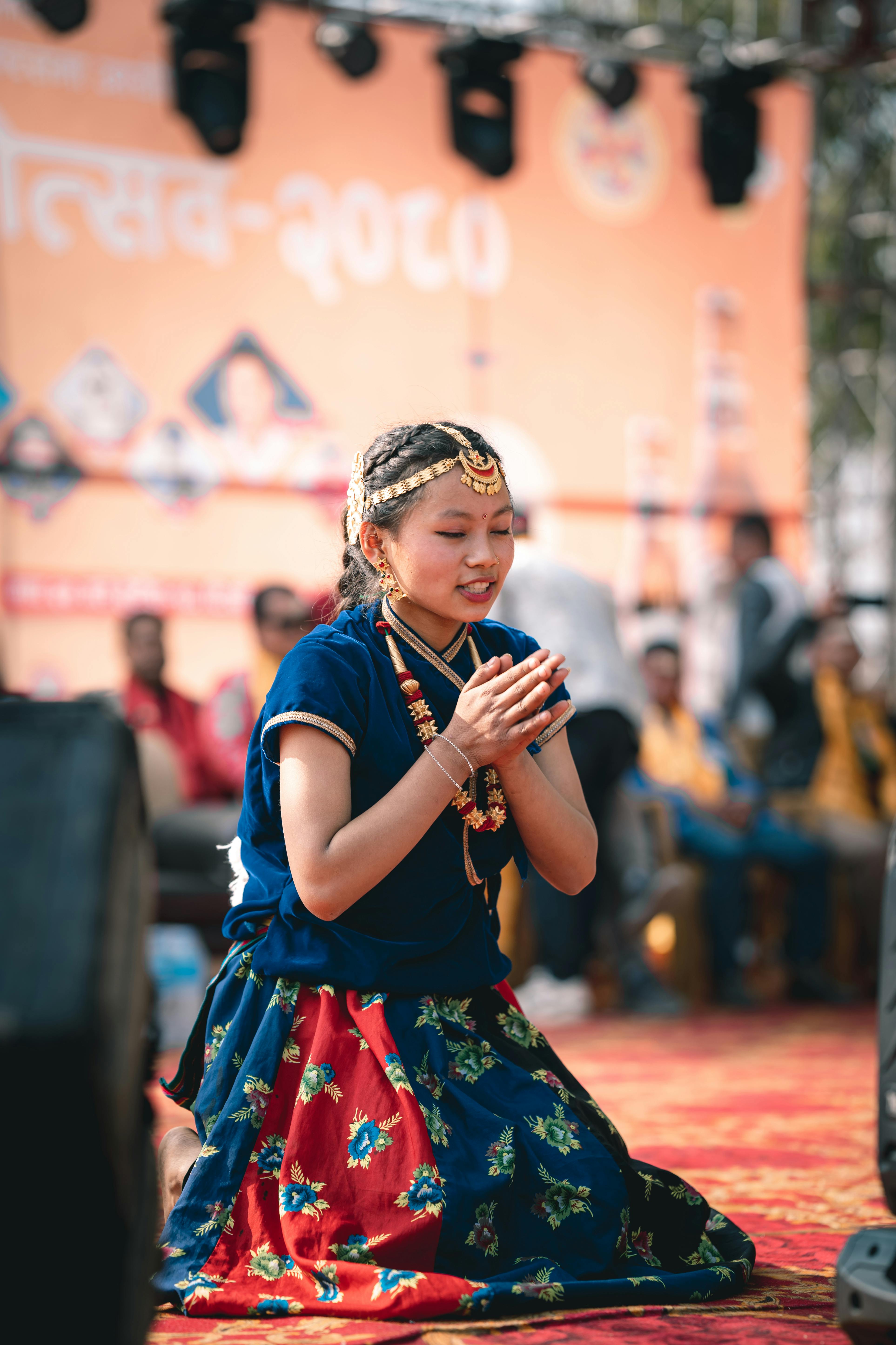 A young woman in traditional attire kneels with clasped hands at a cultural event outdoors.