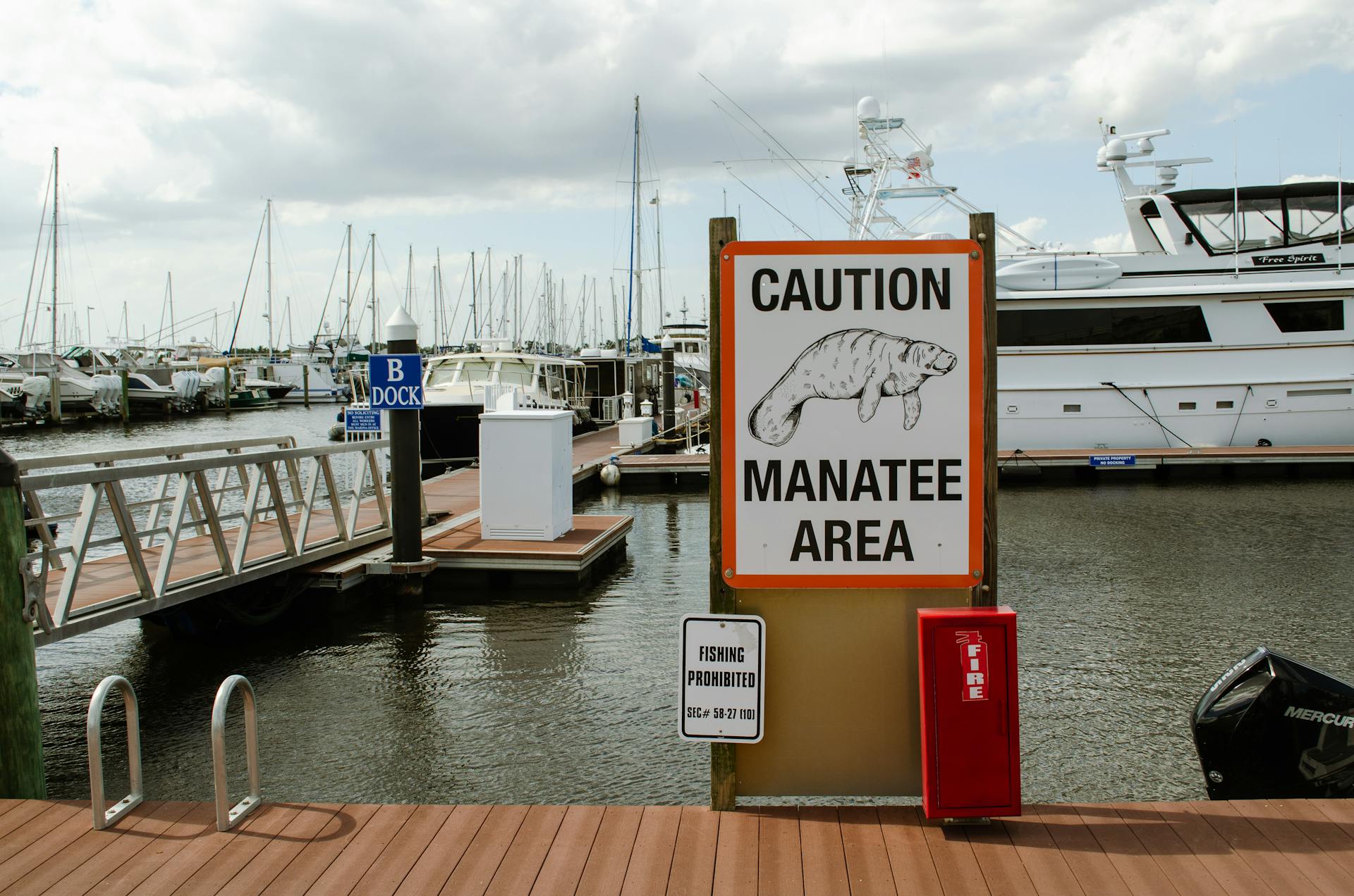 Caution manatee area sign with boats docked at a busy marina under cloudy skies.