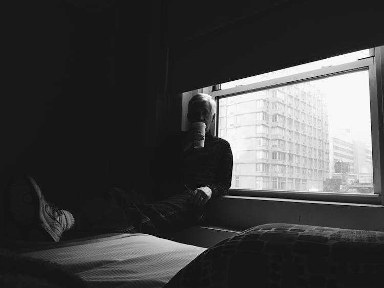 Black And White Photo Of Man Leaning Against Window With Cup Of Coffee