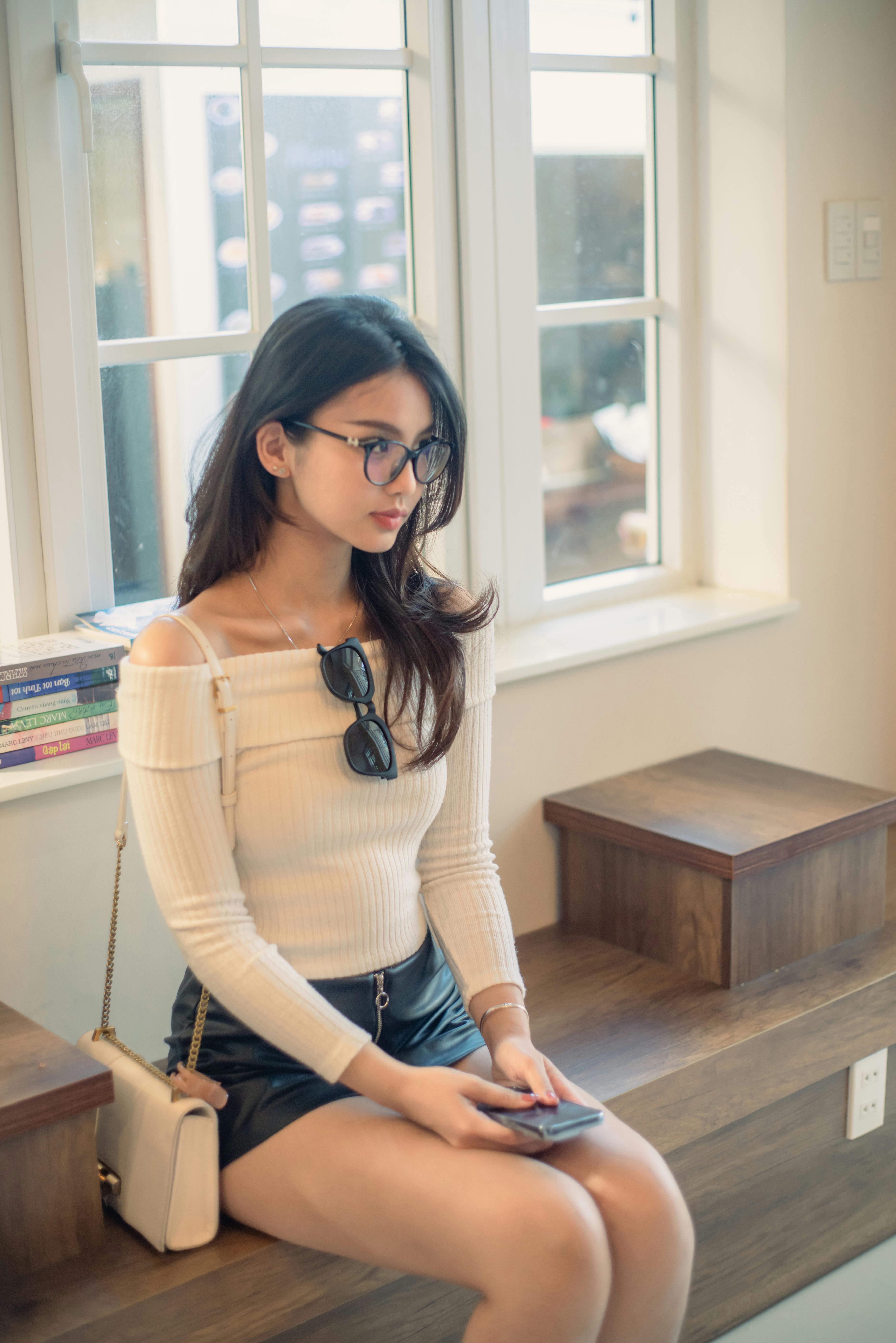 Elegant woman in trendy attire sitting by a window in Dalat, Vietnam, holding her smartphone.