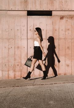 A fashionable woman in high heels walks past a textured wall with a distinct shadow.