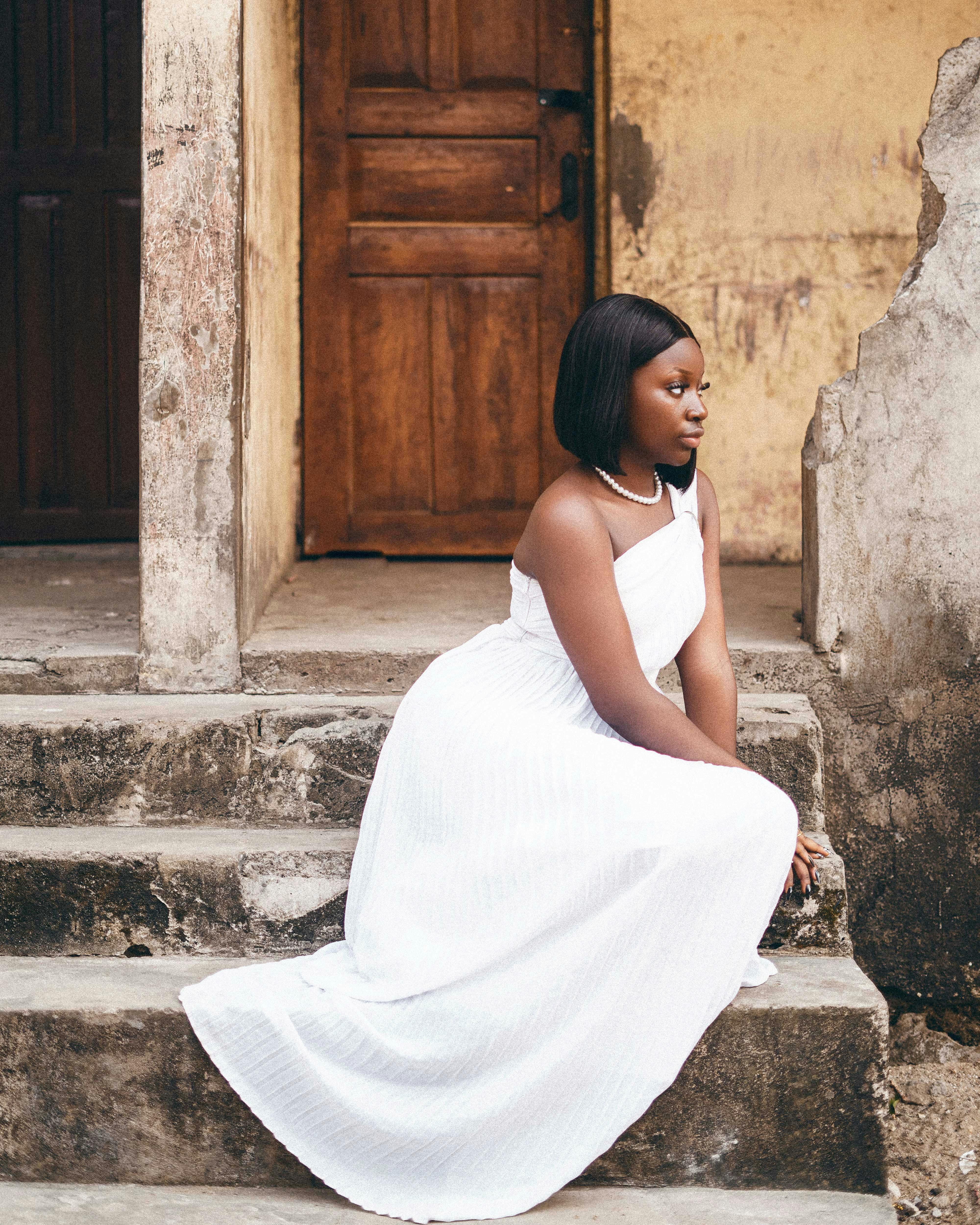 African woman in a white dress poses on rustic steps with wooden doors in background.