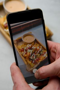 Hand holding smartphone capturing a photo of nachos with cheese and salsa.