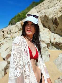 Fashionable woman in red bikini and white tunic poses on rocky beach under bright sun.