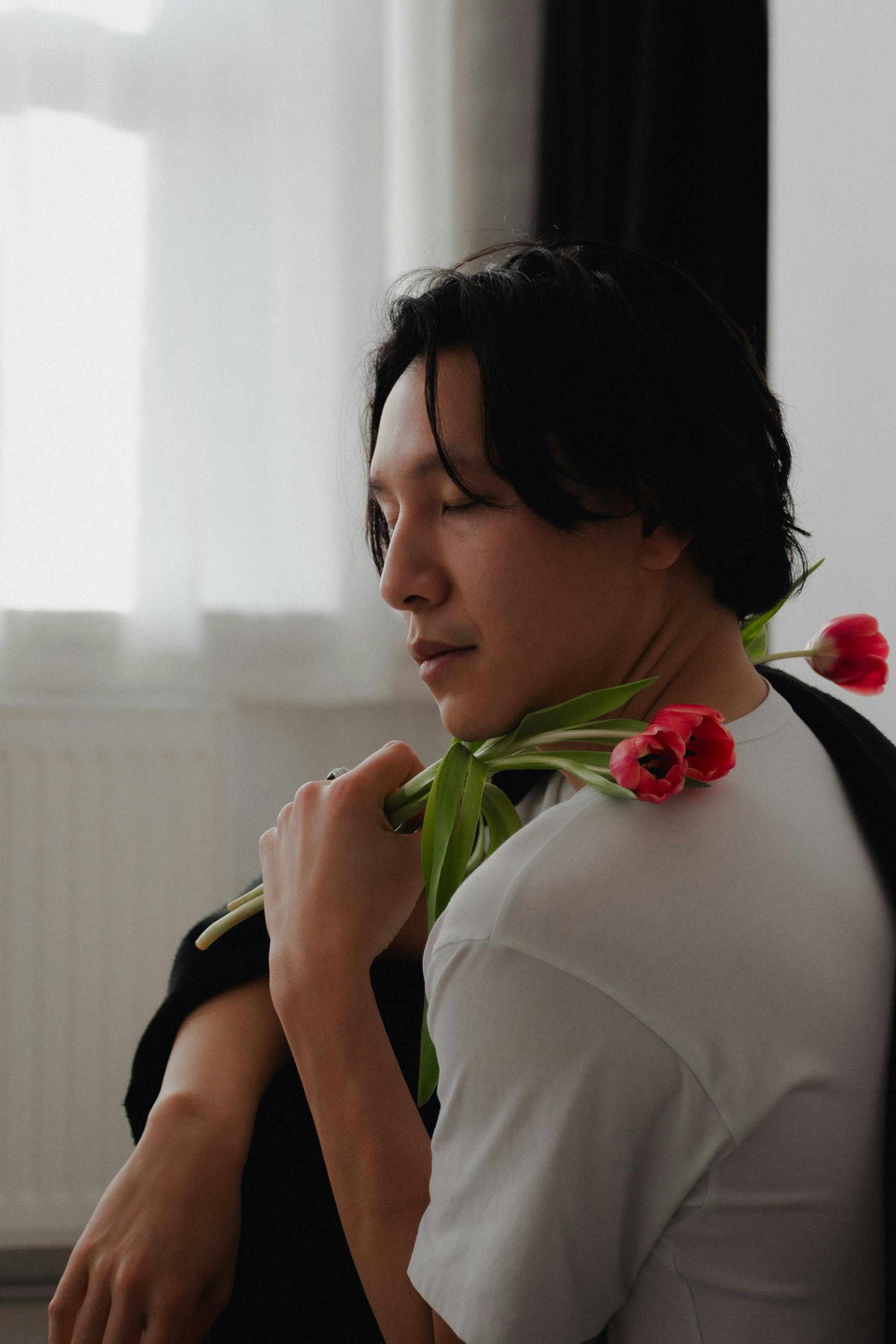 A serene portrait of a man holding red tulips indoors against blurred curtains.