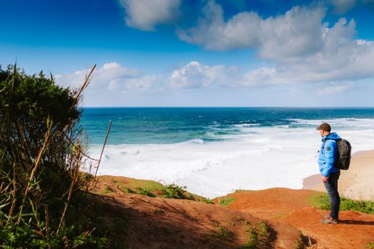 A man in a blue jacket stands on a cliff edge, overlooking the ocean on a sunny day.