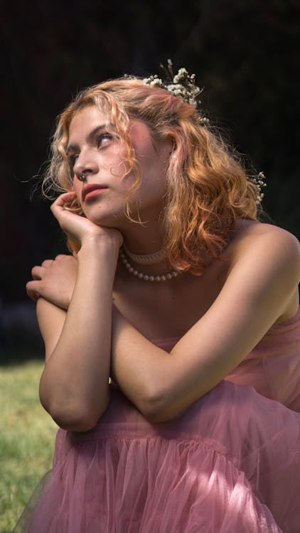 Young Woman in a Pink Dress and Flowers in Hair Sitting in a Garden 